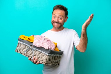 Orta yaşlı bir kadın mavi arka planda izole edilmiş bir çamaşır sepetini tutuyor. Davet edilmesi için ellerini yana uzatıyor.
