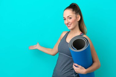 Young sport caucasian woman going to yoga classes while holding a mat isolated on blue background extending hands to the side for inviting to come