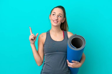 Genç beyaz kadın yoga derslerine giderken mavi arka planda izole edilmiş bir minder tutuyor ve harika bir fikre işaret ediyor.