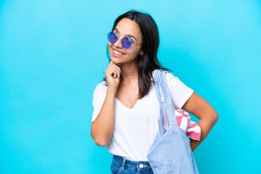 Elinde kumsal çantası olan beyaz bir kadın mavi arka planda izole edilmiş gülümserken yukarı bakıyor.