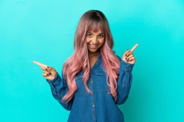 Mavi arka planda izole edilmiş pembe saçlı melez kadın parmak uçlarını işaret ediyor ve mutlu.