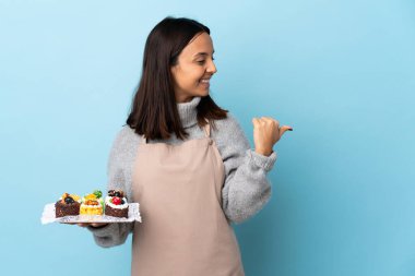 Pasta şefi büyük bir pasta tutuyor mavi arka planda bir ürün sunmak için yan tarafı işaret ediyor.