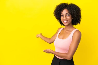 Sarı arka planda izole edilmiş genç Afrikalı Amerikalı kadın davet edilmek için ellerini yana uzatıyor.