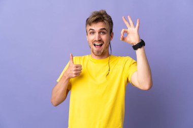Mor arka planda izole edilmiş genç, yakışıklı sarışın adam işaret ve baş parmak işareti gösteriyor.