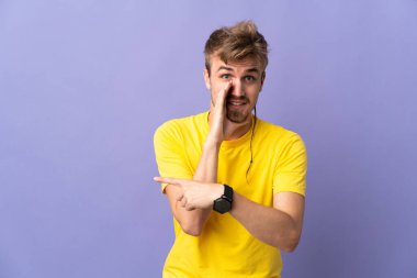 Mor arka planda izole edilmiş genç, yakışıklı sarışın bir adam bir ürün sunmak ve bir şeyler fısıldamak için kenarı işaret ediyor.