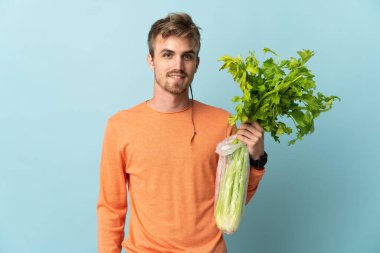Mavi arka planda izole edilmiş bir kereviz tutan genç sarışın adam çok gülümsüyor.