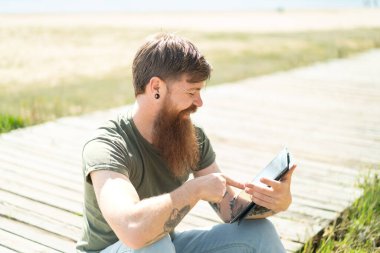 Kızıl saçlı sakallı adam tablet ekranına mutlu bir ifadeyle dokunuyor.