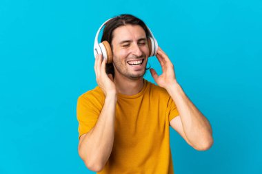 Genç yakışıklı adam mavi arka planda izole edilmiş müzik dinliyor ve şarkı söylüyor.
