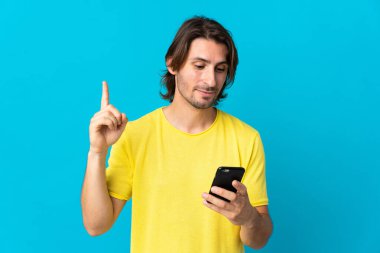 Genç yakışıklı adam mavi arka planda cep telefonu ve parmak kaldırarak izole edilmiş.