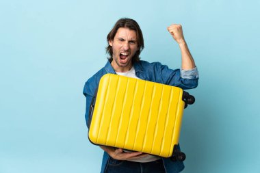 Genç yakışıklı adam mavi arka planda izole edilmiş seyahat valiziyle tatilde.