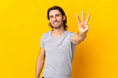 Sarı arka planda izole edilmiş genç yakışıklı mutlu ve parmaklarıyla üçe kadar sayıyor.