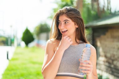 Genç, beyaz, güzel bir kadın elinde bir şişe suyla dışarıda bir fikir üretiyor ve ona bakıyor.