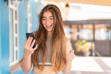 Genç, beyaz bir kadın cep telefonu kullanıyor ve zafer jesti yapıyor.