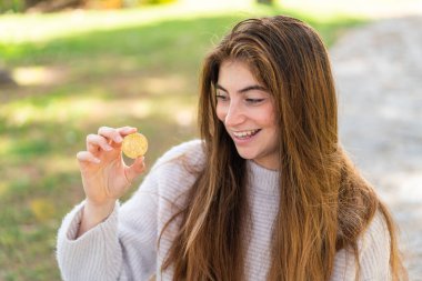 Genç, güzel, beyaz bir kadın elinde Bitcoin ile dışarıda mutlu bir ifadeyle bekliyor.