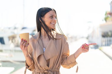 Açık havada elinde kahve tutan genç bir kadın bir ürün sunmak için yan tarafı işaret ediyor.