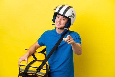 Sırt çantalı genç Brezilyalı adam sarı arka planda izole edilmiş mutlu bir ifadeyle ön tarafı işaret ediyor.