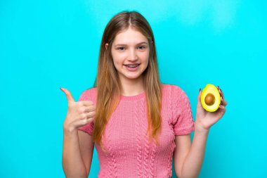 Avokado tutan genç bir Rus kızı mavi arka planda izole edildi. Başparmakları havada çünkü iyi bir şey oldu.
