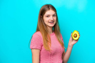 Elinde avokado tutan genç bir Rus kız mavi arka planda izole edilmiş, gülümsüyor.