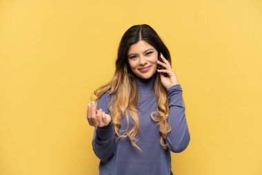 Sarı arka planda izole edilmiş cep telefonu kullanan genç Rus kız para hareketi yapıyor.