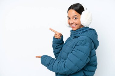 Kış kulaklığı takan genç İspanyol kadın beyaz arka planda izole edilmiş bir şekilde parmak göstererek bir ürün sunuyor.