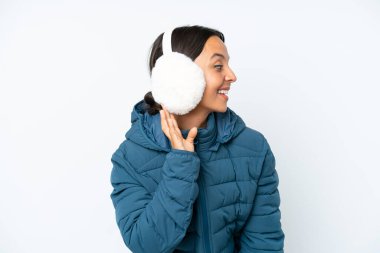 Young hispanic woman wearing a winter earmuffs isolated on white background listening to something by putting hand on the ear