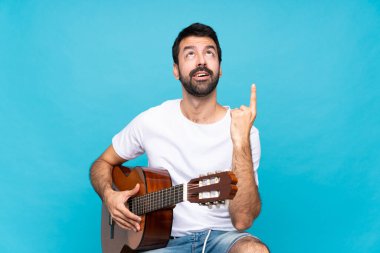 İzole edilmiş mavi arka planda gitar çalan genç adam yukarı bakıyor ve şaşırıyor.