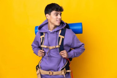 Young mountaineer man with a big backpack over isolated yellow background looking to the side and smiling