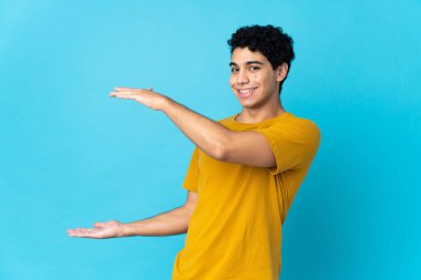 Genç Venezuelalı bir adam mavi arka planda izole edilmiş bir ilan vermek için kopyalama alanı tutuyor