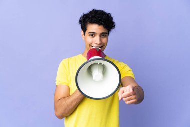 Genç Venezuelalı bir adam mor arka planda izole edilmiş bir şekilde megafondan bağırarak bir şeyi duyurmak için ön tarafı işaret ediyor.