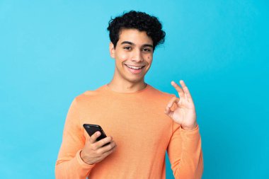 Venezuelalı adam mavi arka planda cep telefonu kullanarak izole edilmiş ve tamam işareti yapıyor.