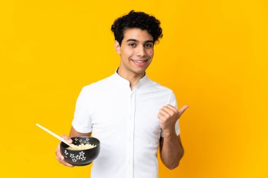 Genç Venezuelalı, sarı arka planda izole edilmiş bir adam elinde çubuklarla bir kase erişte tutarken bir ürünü sunmak için yan tarafı işaret ediyor.