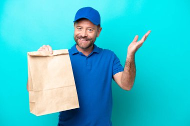 Orta yaşlı bir adam mavi arka planda izole edilmiş bir paket yiyecek alıyor. Davet için ellerini yana uzatıyor.