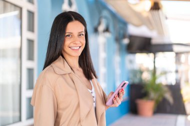 Açık havada cep telefonu kullanan genç bir kadın. Çok gülümsüyor.