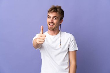 Genç yakışıklı sarışın adam mor arka planda izole edildi. Başparmakları havada, çünkü iyi bir şey oldu.