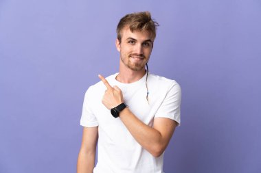 Mor arka planda izole edilmiş yakışıklı sarışın bir adam bir ürün sunmak için kenarı işaret ediyor.