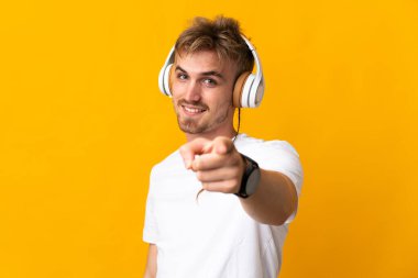 Sarışın genç adam sarı arka planda izole edilmiş müzik dinliyor ve ön tarafı işaret ediyor.