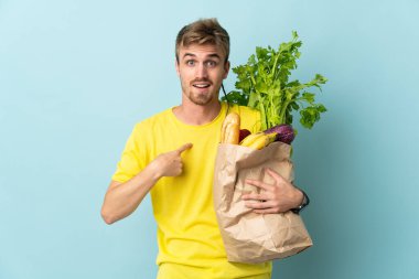 Sarışın kişi, mavi arka planda izole edilmiş bir paket yemek alıyor. Şaşırtıcı yüz ifadesi var.