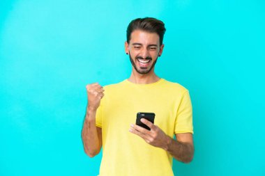 Kafkasyalı genç adam mavi arka planda izole edilmiş ve telefon zafer pozisyonunda.