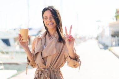 Açık havada elinde kahveyle gezen genç bir kadın gülümsüyor ve zafer işareti gösteriyor.