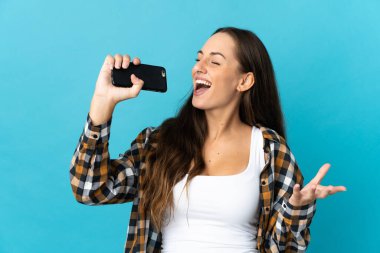 İzole edilmiş mavi arka planda cep telefonu kullanan ve şarkı söyleyen genç İspanyol kadın.