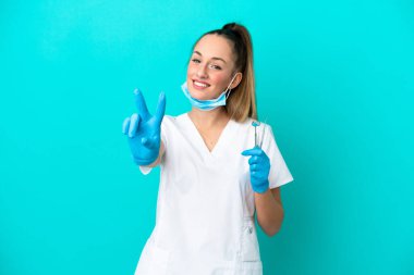Dişçi beyaz kadın, elinde mavi arka planda izole edilmiş aletler tutuyor. Gülümsüyor ve zafer işareti gösteriyor.