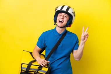 Sırt çantalı genç Brezilyalı adam sarı arka planda izole edilmiş gülümsüyor ve zafer işareti gösteriyor.