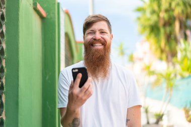 Kızıl saçlı, sakallı, açık havada cep telefonu kullanan, gülümseyen bir adam.