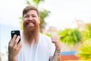 Kızıl saçlı, sakallı, cep telefonu kullanan ve yan tarafı işaret eden adam.