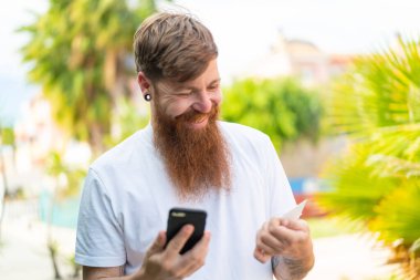 Sakallı kızıl saçlı adam kredi kartıyla cep telefonu alıyor.