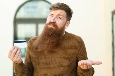 Sakallı kızıl saçlı adam dışarıda kredi kartı tutuyor. Omuzlarını kaldırırken şüpheyle hareket ediyor.