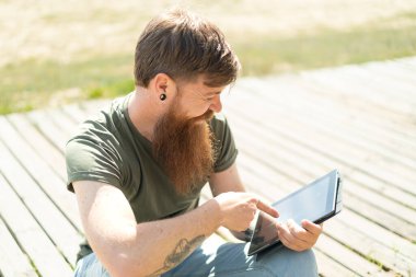 Kızıl saçlı sakallı adam tablet ekranına mutlu bir ifadeyle dokunuyor.