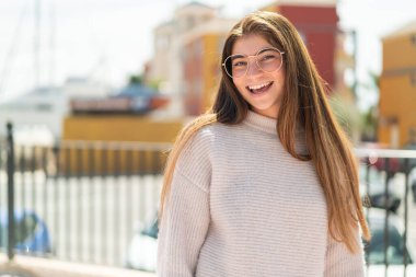 Açık havada gözlüklü, güzel, beyaz bir kadın. Yüzünde sürpriz bir ifade var.