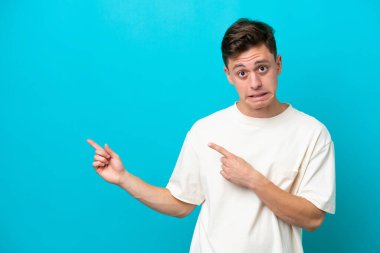 Genç ve yakışıklı Brezilyalı adam mavi arka planda izole edilmiş korkmuş ve kenarı işaret ediyor.