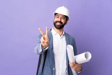 Miğferli genç mimar, izole edilmiş mor arkaplanının planlarını tutuyor. Gülümsüyor ve zafer işareti gösteriyor.